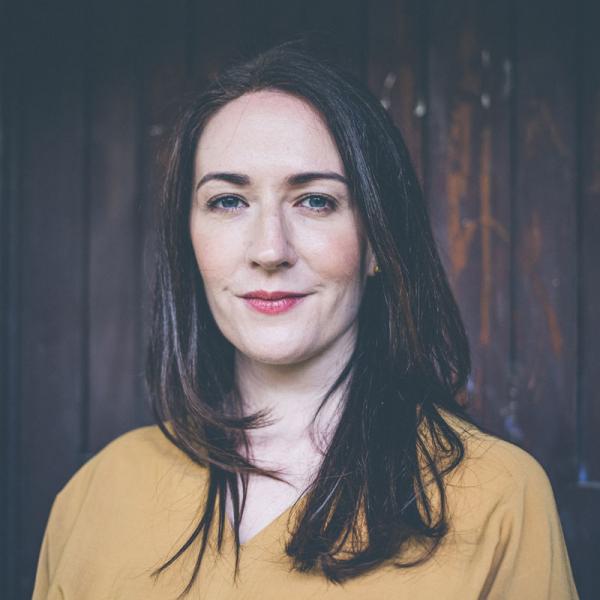 Headshot photo Peggy Nolan wearing a mustard yellow top against a dark background
