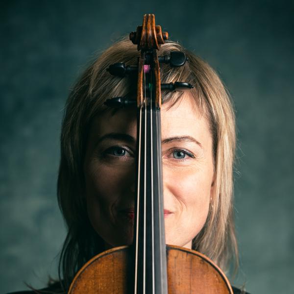 Ioana Petcu-Colan smiles at the camera from behind the neck of a violin, which perfectly disects her face.  Violin 