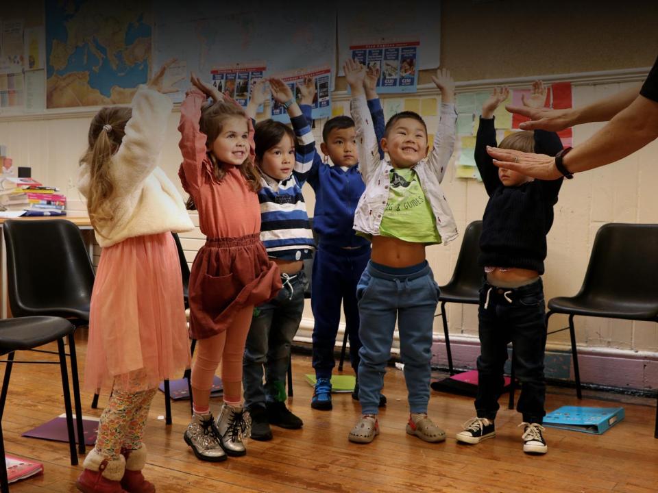 A group of smiling 4 - 5 yar old boys and girls hold their arms in the air as they play a game based on music and learning