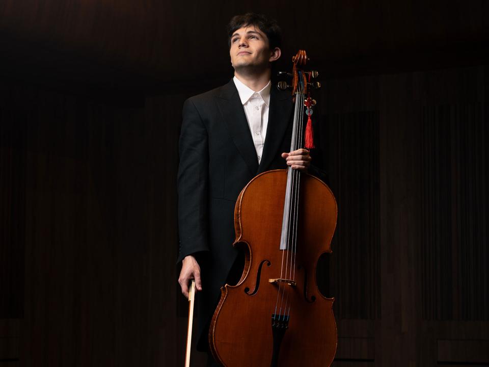 Andrés Vacarro stands in a beam of light holding a cello and bow
