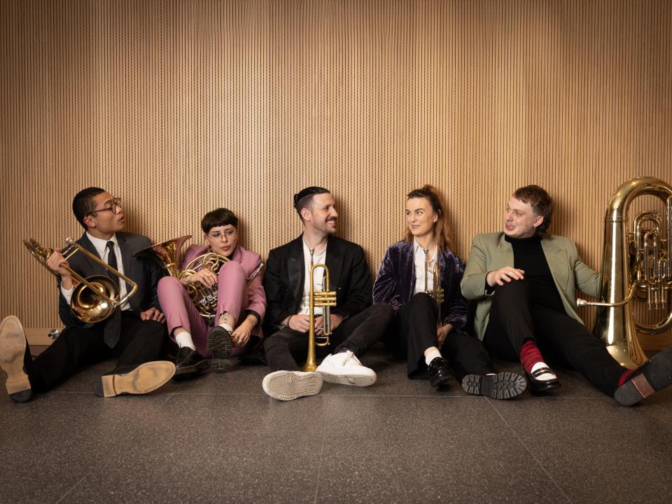 Members of Banchee Brass Quintet in smart causal clothes sit ont he floor while holding their instruments and laughing 