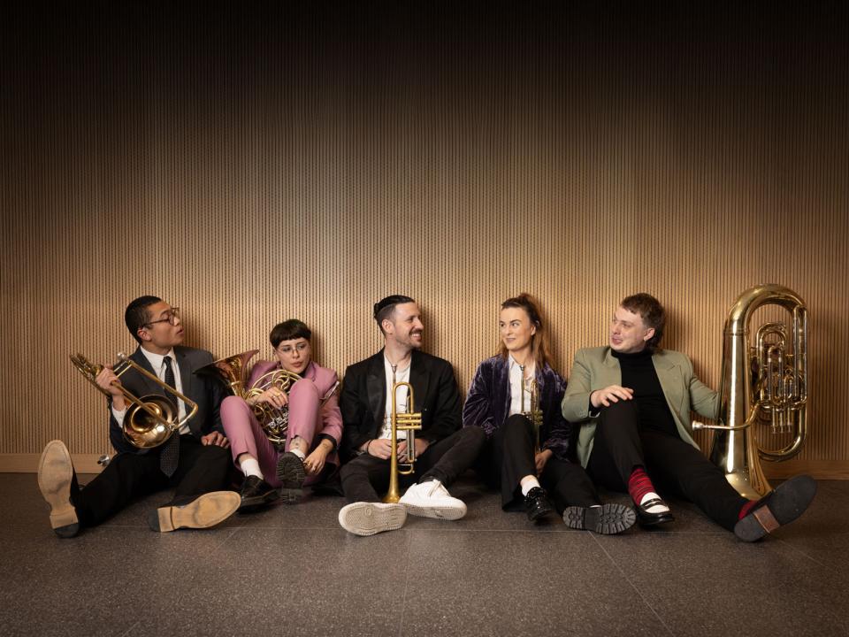 A brass quartet of young mixed gender students sit on a terrao floor with the instruments leaning against light-coloured wood panelling. 