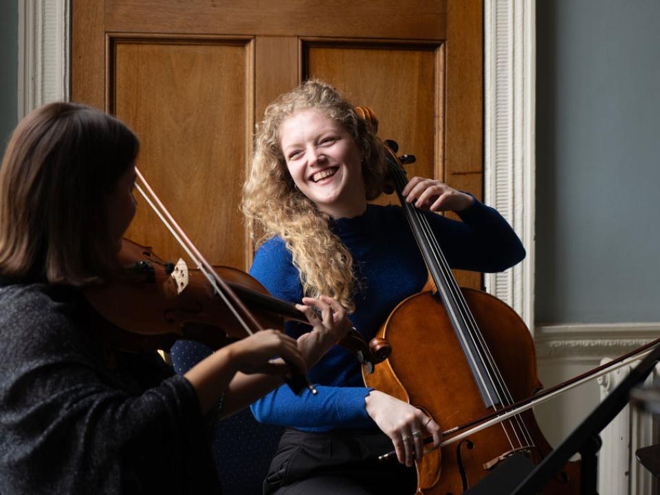 A student with long culrey blonde hair plays cello and laughs with another musician who's face we cannot see