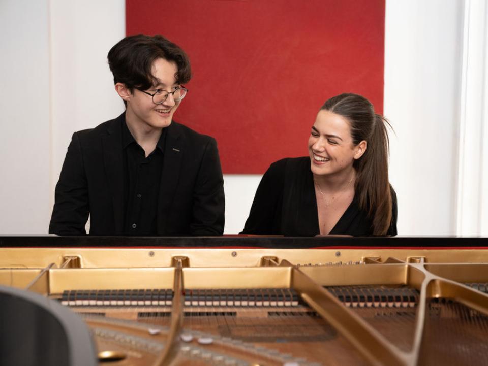 Two smiling students sit behind a piano enjoying a moment of lightheartedness during a rehearsal 