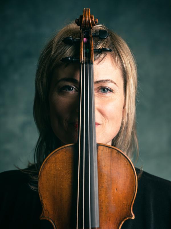 Ioana Petcu-Colan smiles at the camera from behind the neck of a violin, which perfectly disects her face.  Violin 