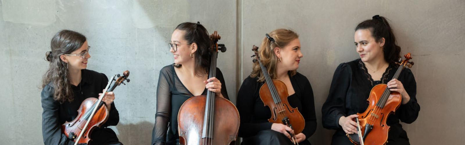 Four classical string musicians in black concert dress, holding two violins, a viola and a cello, sit on wooden benches against a light concrete wall, smiling and talking together.