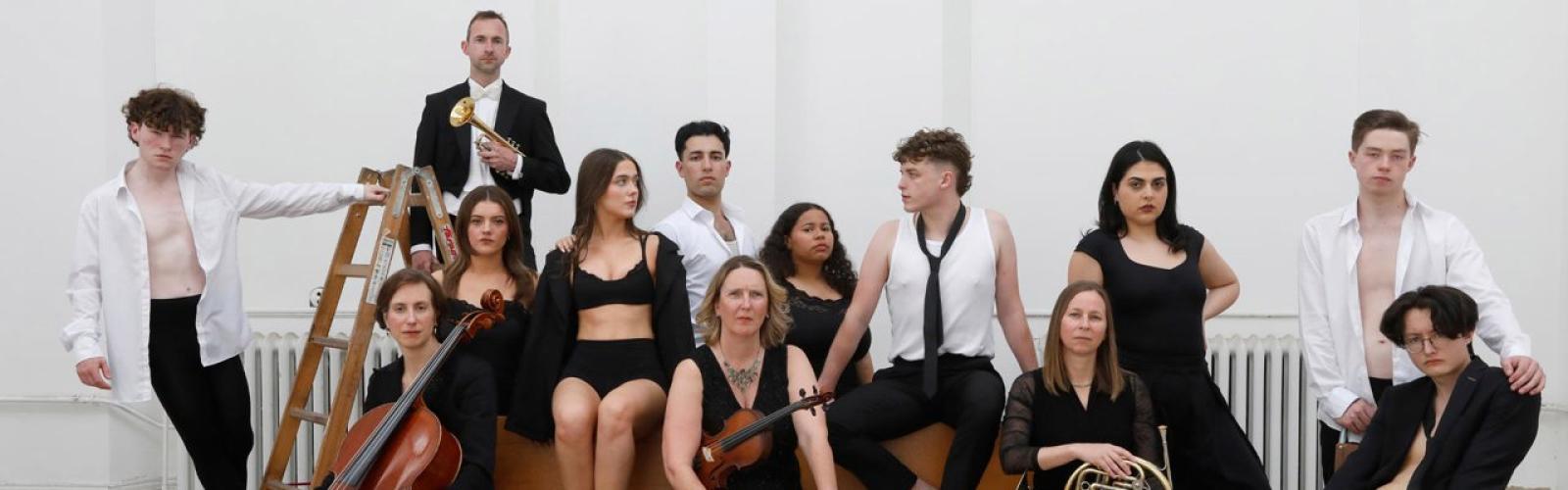 A group of young musical theatre performers sit and stand alongside orchestral musicians holding instruments, including a cello, violin, french horn and trumpet. The musicians are wearing formal concert blacks while the cast members wear casual black and white clothing. 