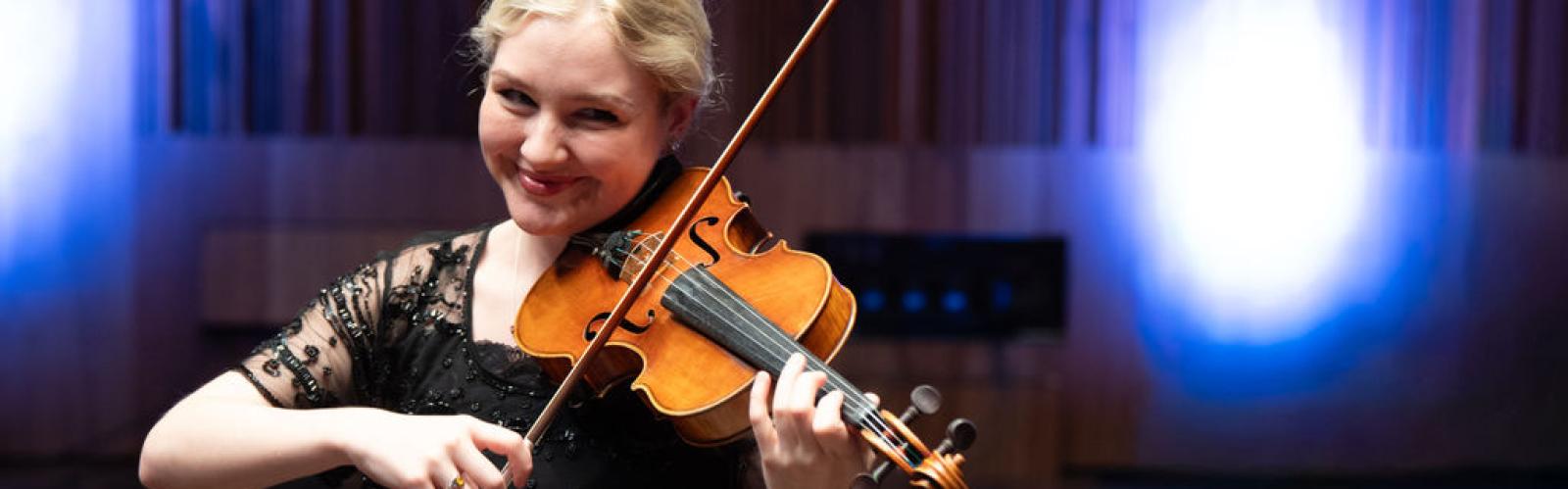 A young adult with blonde hair performs the violin in front of bright blue stage lights 