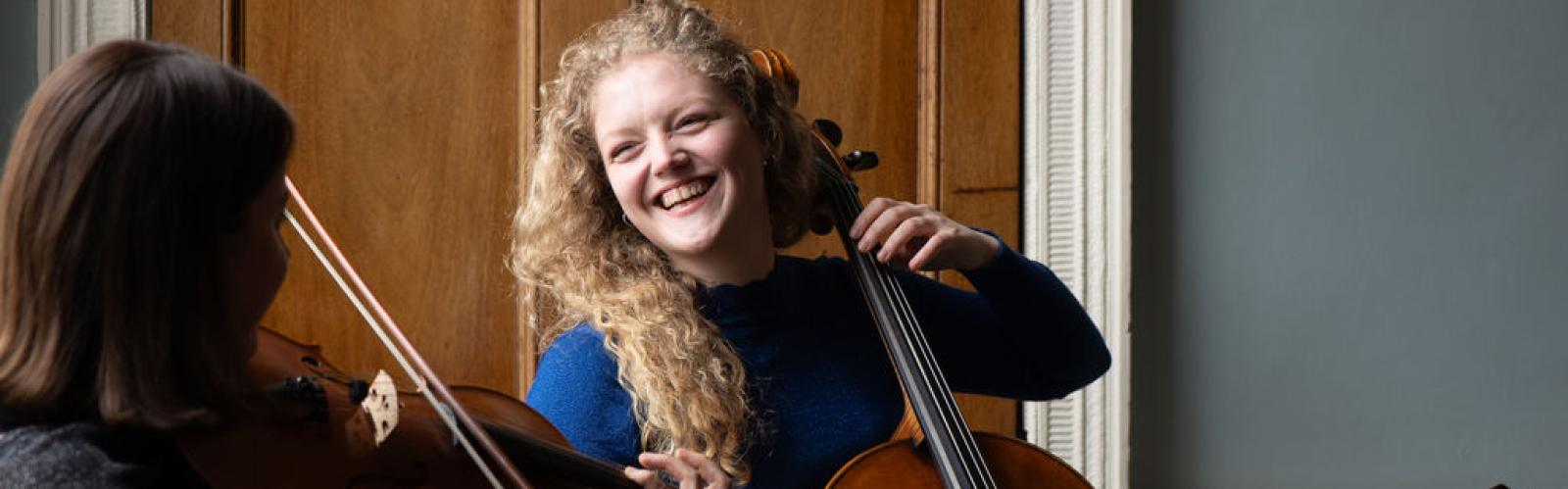 A student with long culrey blonde hair plays cello and laughs with another musician who's face we cannot see